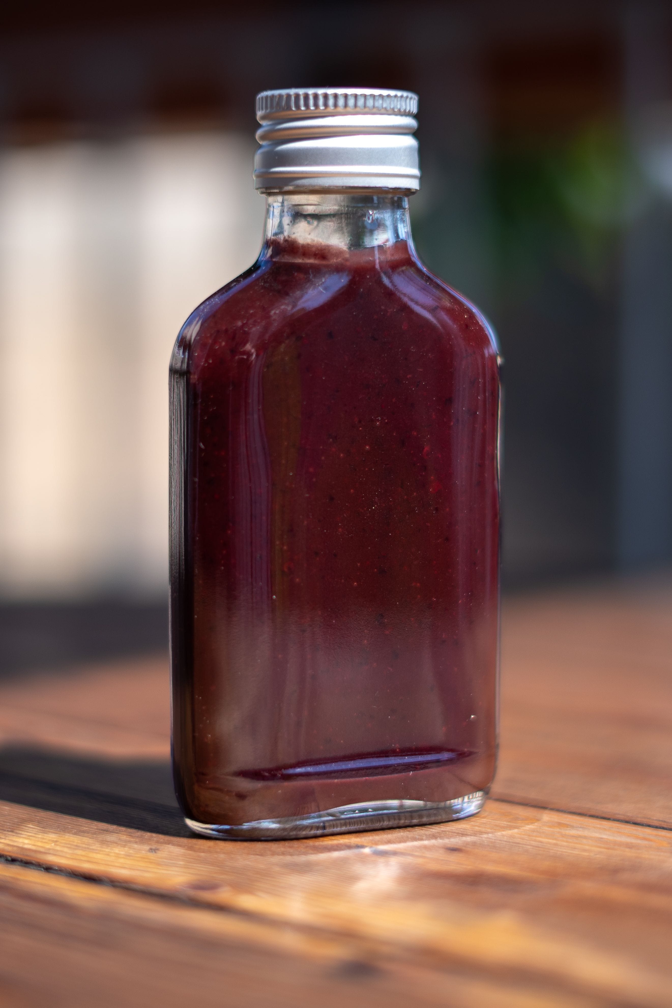 The image shows a small, clear glass bottle with a silver screw cap, filled with a dark, thick sauce. The sauce is deep violet, with a slightly glossy texture. The bottle is placed on a wooden surface, with a blurred background.