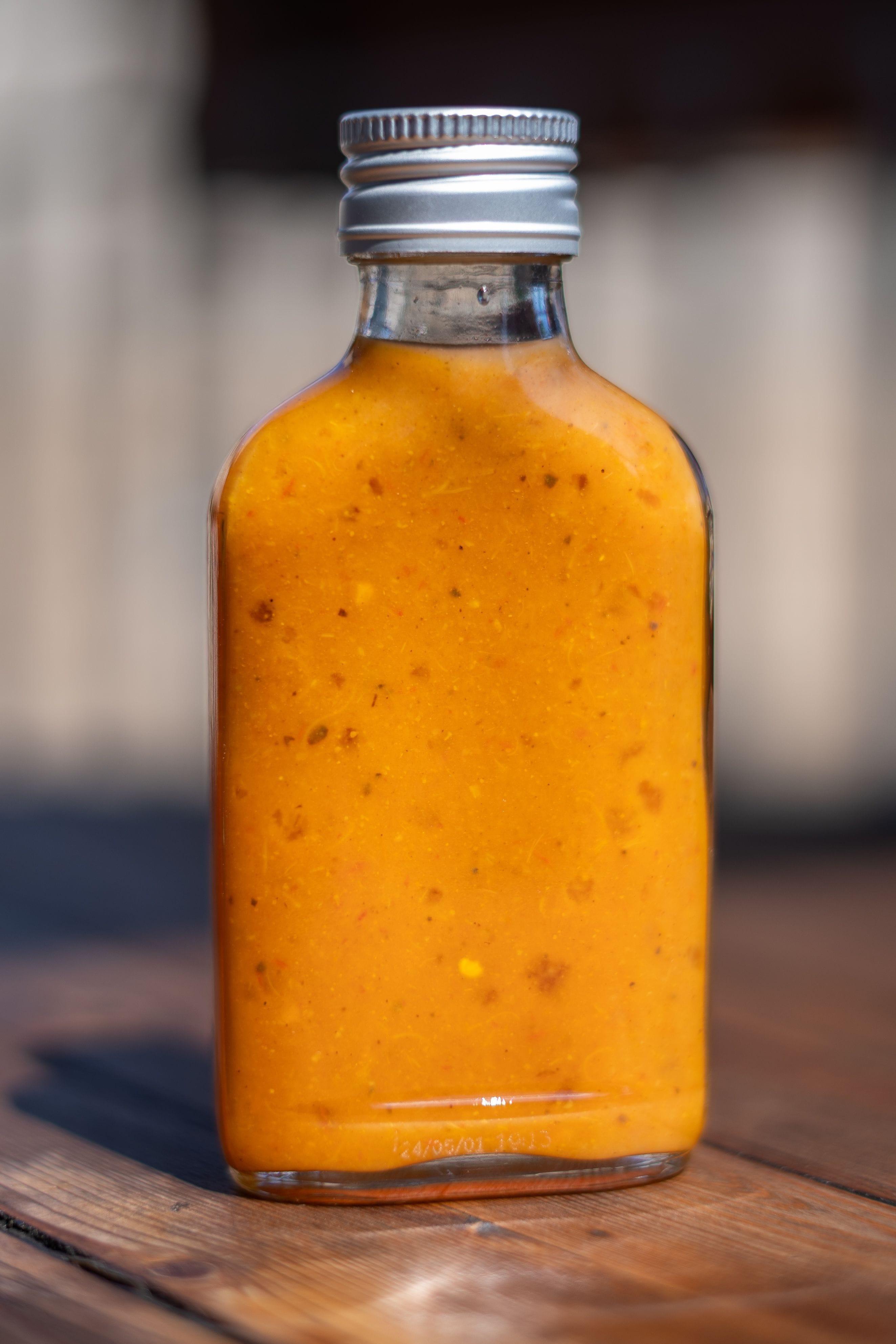 The image shows a small, clear glass bottle filled with a thick, orange-colored sauce. The bottle has a silver metal screw cap. The sauce appears to have small specks of spices or ingredients, giving it a textured appearance. The bottle is placed on a wooden surface.