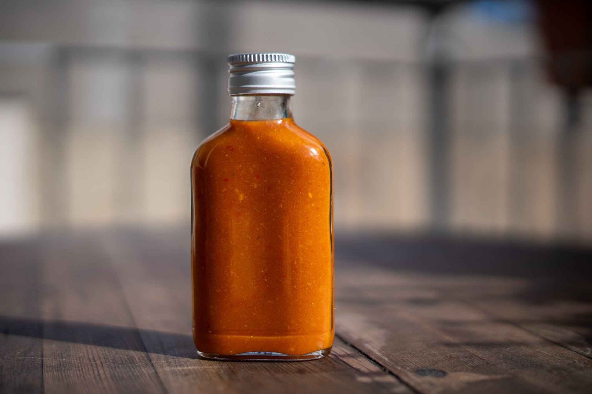 A small glass bottle filled with orange-colored hot sauce, sits on a wooden surface. The bottle has a silver metal cap, and the background is softly blurred, drawing focus to the bottle. The lighting highlights the texture of the sauce inside.