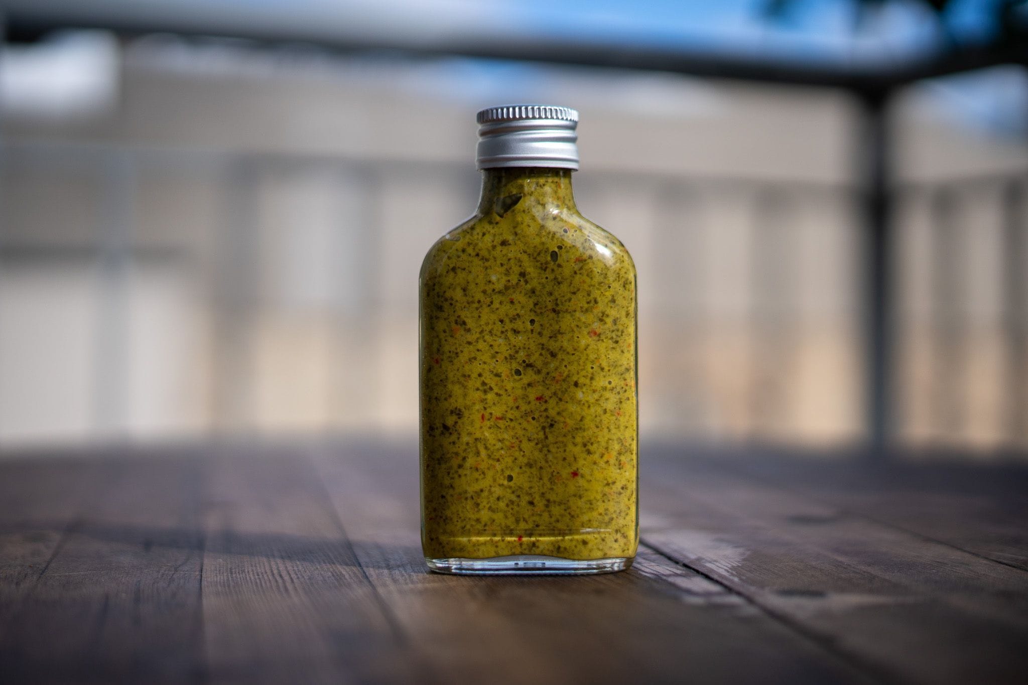 A small glass bottle with a silver cap is filled with a thick, greenish-yellow sauce or dressing, speckled with herbs and spices. The bottle is placed on a wooden surface with a blurred background, possibly outdoors, under natural light.