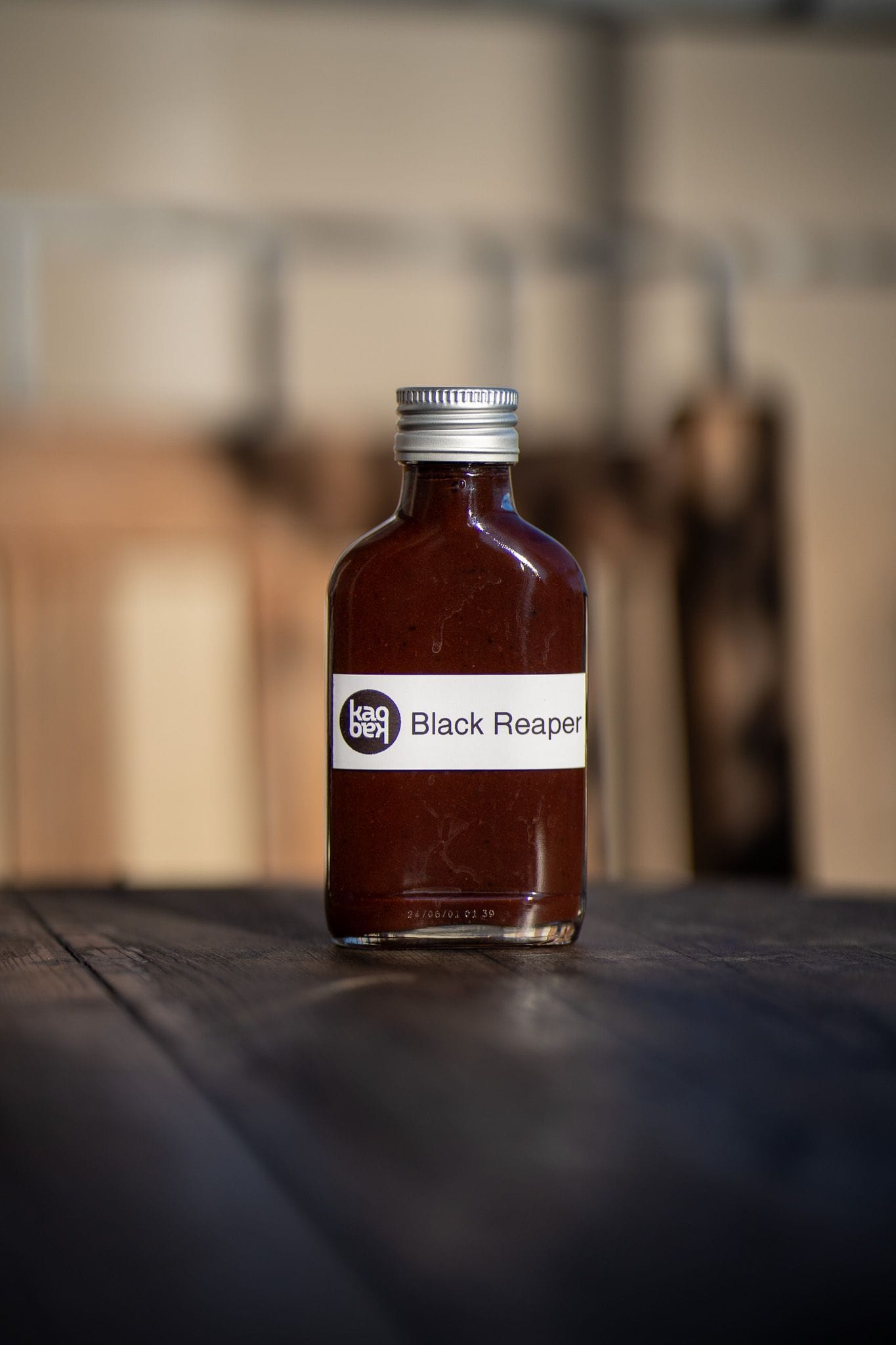 A small glass bottle with a silver cap, filled with a dark brown liquid. The label reads "Black Reaper" with a logo. The bottle is placed on a wooden surface with a blurred background, giving a warm and rustic ambiance.